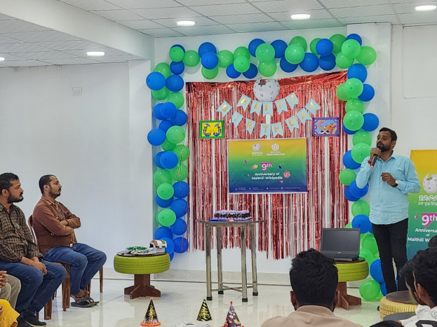 Eine Gruppe von Menschen sitzt auf Stühlen vor einer Bühne mit Ballons, einem Banner, einer Uhr und anderen Gegenständen, während eine Person auf der Bühne ein Mikrofon in der Nähe eines Tisches mit einem Laptop und anderen Gegenständen hält, unter Deckenlampen für eine 9. Geburtstagsfeier.