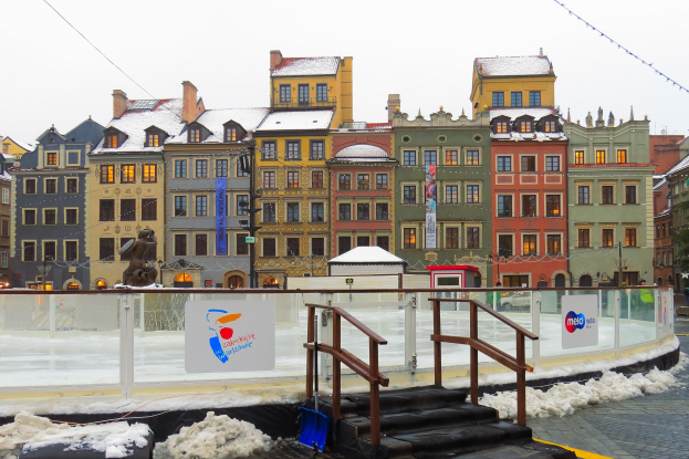 Eisbahn in einem Stadtplatz umgeben von Geb├Ąuden, Treppen, Gel├Ąndern, Informationsschildern, schneebedeckter Erde, Laternenmasten, B├Ąumen und Himmel.
