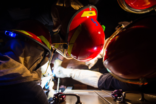 Eine Gruppe von Feuerwehrleuten in Helmen und Handschuhen arbeitet an einem Feuerwehrauto vor einem dunklen Hintergrund, mit einem Tisch voller verschiedener Gegenstände auf der rechten Seite.