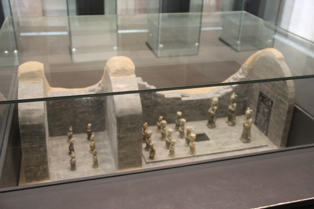 Ein Museumsvitrine mit verschiedenen Artefakten, darunter Skulpturen und Glasboxen, vor einer Wand.