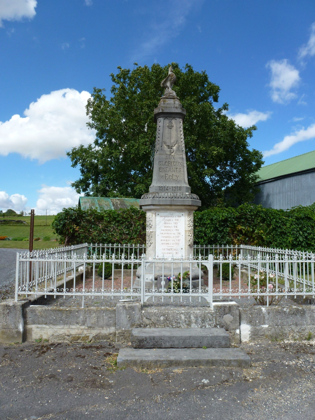 Kriegsdenkmal mit eingravierter Inschrift, umgeben von einem Metallzaun, Treppe führt hinauf, in einem Friedhof mit Pflanzen, Bäumen, einer Hütte und bewölktem Himmel.
