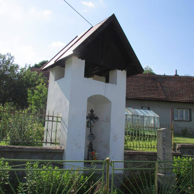 Kleines weißes Kirchlein mit einem Kreuz darauf, umgeben von einem Metallzaun, vor einem bewölkten Himmel, umgeben von Pflanzen, einem Weg, Häusern, Pfählen und Drähten.