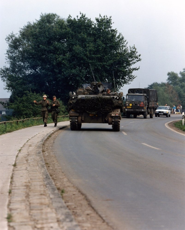 Ein militärisches Fahrzeug fährt eine Straße neben einem Wald entlang, während uniformierte Personen daneben gehen und Gebäude sowie ein klarer blauer Himmel im Hintergrund zu sehen sind.