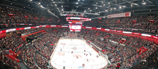 Eine große, hell erleuchtete Arena voller Zuschauer, die während eines Eishockeyspiels jubeln, mit einem zentralen Bildschirm über der Eisfläche.