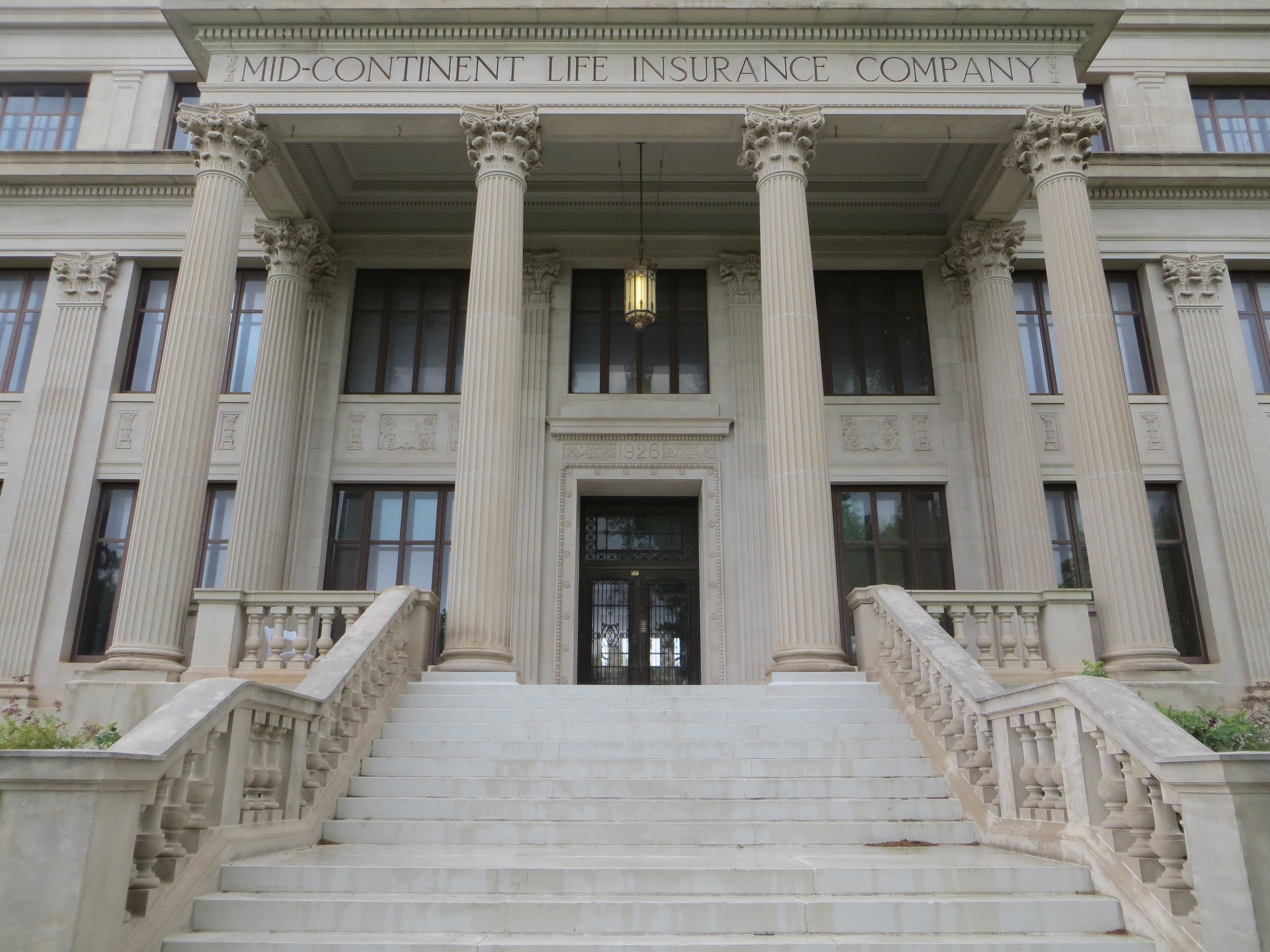 Großes Gebäude mit der Aufschrift "Mid-Continental Life Insurance Company", Fenstern, Säulen, einer Tür, einer Treppe mit Geländer und umliegenden Pflanzen.