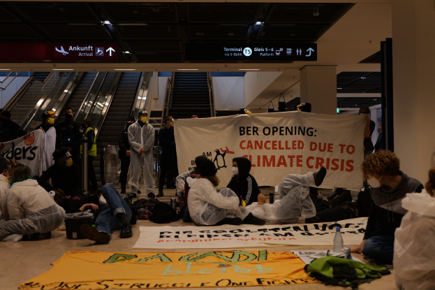 Eine Gruppe von Menschen sitzt auf dem Boden eines Flughafens und hält ein Banner mit der Aufschrift "Berlin wegen Klimakrise abgesagt", mit verstreuten Taschen, Flaschen und anderen Gegenständen und Rolltreppen, Texttafeln und Deckenleuchten im Hintergrund.