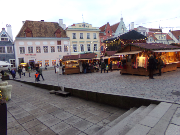 Ein geschäftiger Weihnachtsmarkt in Tallinn, Estland, mit Menschen um dekorierte Stände, festliche Lichter und Topfpflanzen, vor dem Hintergrund bewölkter Himmel und beleuchteter Fenster.