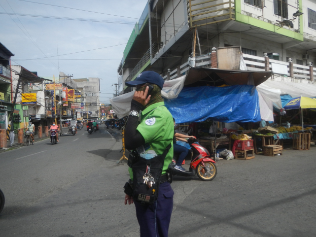Ein Polizist in grüner und blauer Uniform und Mütze steht auf einer Straße und spricht in ein Handy. Im Hintergrund sind Fahrzeuge und Marktstände zu sehen sowie ein Gebäude mit Wolken am Himmel.