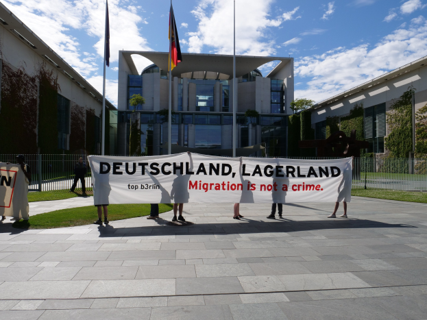 Gruppe von Menschen mit einem Banner "Deutschland, Lagerland Migration ist kein Verbrechen" vor einem Gebäude mit Fenstern, umgeben von Gras, einem Metallzaun, Pflanzen, Bäumen und Fahnen mit Stangen unter einem bewölkten Himmel.
