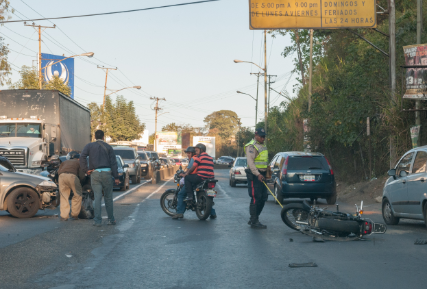 Eine Gruppe von Menschen umringt ein verunglücktes Motorrad am Straßenrand mit mehreren Fahrzeugen, darunter ein Lastwagen, und einer Hintergrundlandschaft aus Bäumen, Pfosten, Lampen und Schildern unter dem Himmel.