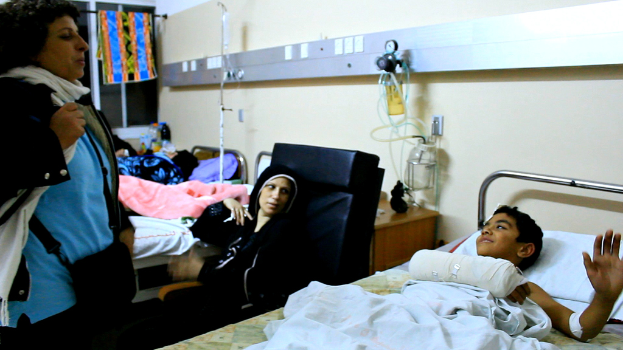 Ein Blick ins Innere eines Krankenhauses mit Betten und Patienten, Kochsalzflaschen auf Ständern zwischen den Betten und einer Frau mit einer Tasche in der Nähe eines Patienten.
