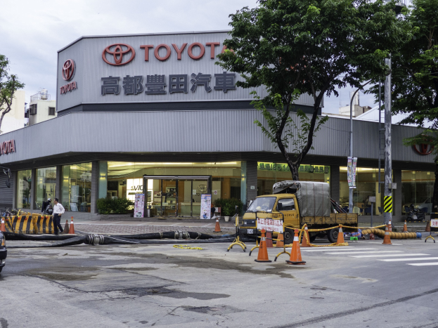 Ein Toyota-Autohaus in Tokio, Japan, mit Fahrzeugen, Verkehrskegeln, Rohren, Menschen, Bäumen, Polen, Brettern, Gebäuden und einer bewölkten Himmel.