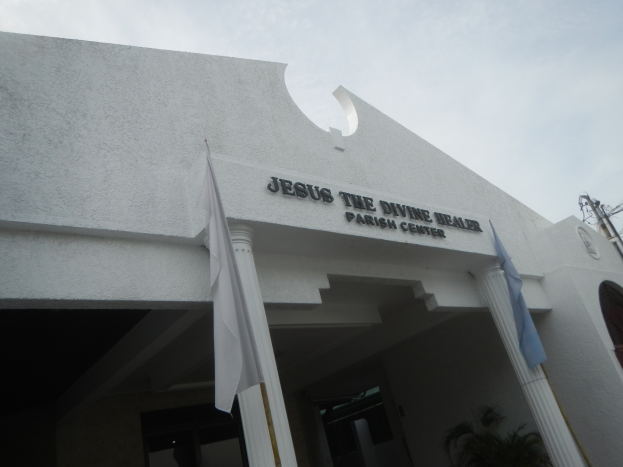 Das Jesus der göttliche Heiler Pfarrzentrum mit Text an der Wand, Säulen, Fahnen an Stangen, einer Pflanze, einem Strommast mit Drähten und einem bewölkten Himmel im Hintergrund.