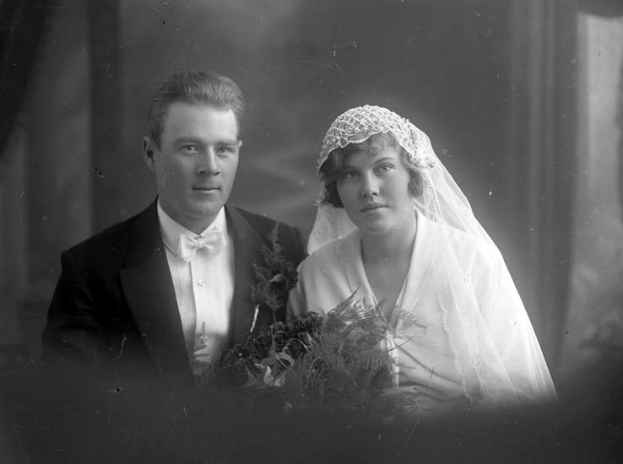 Ein Schwarz-Weiß-Foto eines Mannes in einem Anzug und einer Frau mit einem Blumenstrauß an ihrem Hochzeitstag, mit einem unscharfen Hintergrund.