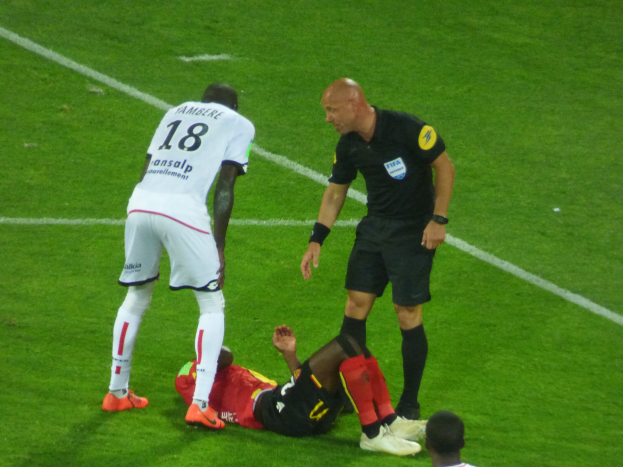 Ein Fußballspieler liegt neben einem Schiedsrichter auf dem Boden, umgeben von drei stehenden Personen auf einem Rasenfeld, wobei eine Person in der Mitte mit entschlossener Miene und ausgestreckten Armen zu sehen ist.
