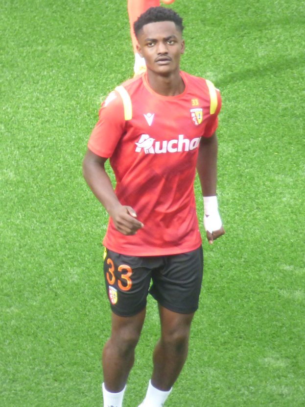 Emmanuel Adebayor, der ein rotes Hemd und schwarze Shorts trägt, geht auf einem Fuballfeld mit zwei Personen im Hintergrund.