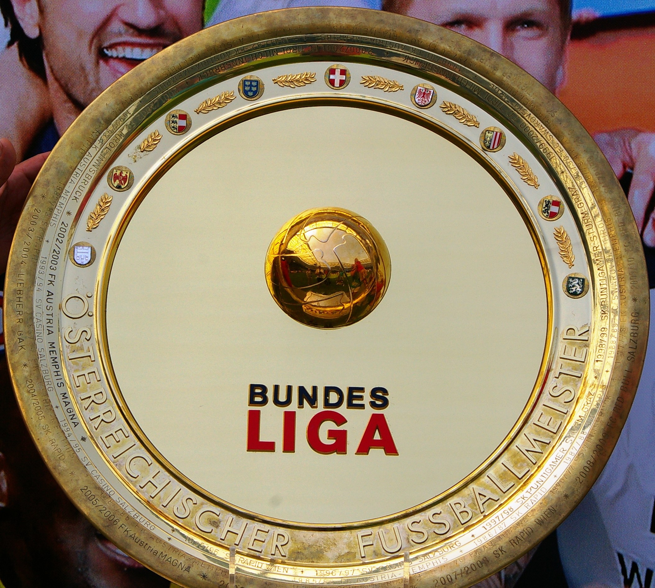 Ein Mann hält einen goldenen Schild mit "Bundesliga" darauf, mit einem Banner im Hintergrund mit drei Personen.