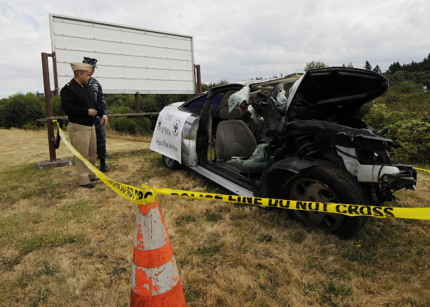 Ein Polizeibeamter in Uniform steht neben einem schwer beschädigten Auto am Straßenrand, mit eingedellter Haube und zersplitterten Fenstern, neben einem Verkehrskegel und einem Band an einem Pfahl, mit Bäumen und einem bewölkten Himmel im Hintergrund.
