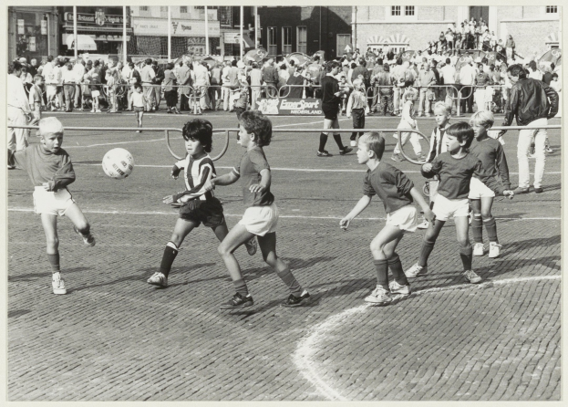 Eine Gruppe junger Jungs, die auf der Straße Fußball spielen, mit einem Ball in der Mitte des Geschehens, in Schwarz-Weiß. Im Hintergrund stehen Menschen hinter einem Geländer, mit Gebäuden mit Fenstern und Pfählen.