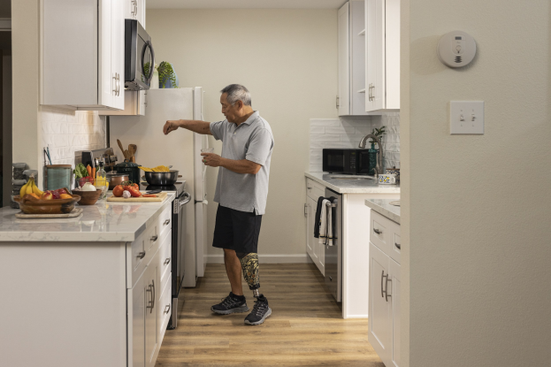 Ein Mann steht in einer Küche mit weißen Schränken und hält ein Glas in der Hand, während verschiedene Gegenstände, einschließlich Obst, Gemüse, Schüsseln und Löffel, auf einer Arbeitsplatte zu seiner Linken liegen.
