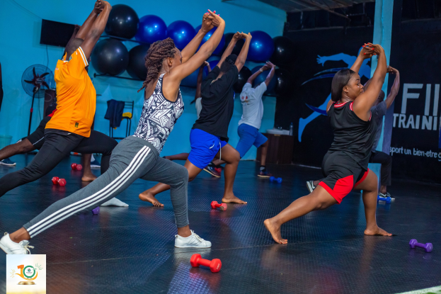 Gruppe von Menschen, die Yoga in einem Fitnessstudio praktizieren, mit Hanteln auf dem Boden, einem Banner mit der Aufschrift "Fitness-Training in Ghana" im Hintergrund und verstreuten Fitnessgeräten.