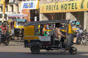 Eine belebte Straße mit verschiedenen Fahrzeugen, darunter ein Auto-Rikscha, und Menschen mit Helmen, mit einer Ampel im Vordergrund und Gebäuden mit Namensschildern und Rollläden im Hintergrund.
