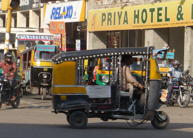 Eine belebte Straße mit verschiedenen Fahrzeugen, darunter ein Auto-Rikscha, und Menschen mit Helmen, mit einer Ampel im Vordergrund und Gebäuden mit Namensschildern und Rollläden im Hintergrund.