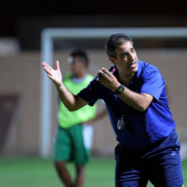Ein Fußballtrainer in einem blauen T-Shirt und Hose steht auf einem Feld und hält ein Mikrofon in der Hand, während er auf Spieler zeigt. Im Hintergrund ist ein Torpfosten und eine Person in einem grünen Kleid zu sehen.