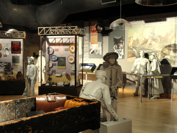 Ein Museumsraum mit verschiedenen Exponaten, darunter Puppen mit H├╝ten, einer Aktentasche, einer Kiste und anderen Gegenst├Ąnden auf dem Boden, mit Informationsschildern an den W├Ąnden und Lampen an der Decke.