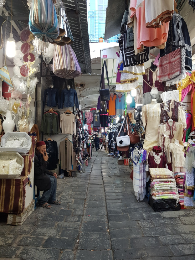 Ein belebter Straßenmarkt in der Altstadt von Jerusalem mit Ständen, die Kleidung, Taschen, Lampen und andere Artikel verkaufen, Menschen, die durch die Gassen schlendern, und ein Gebäude im Hintergrund.