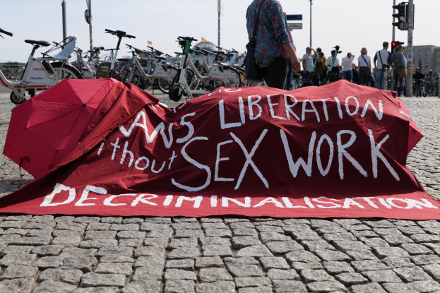 Eine Gruppe von Menschen, die auf dem Boden stehen, einige halten Schirme, mit einem roten Tuch mit der Aufschrift "Befreiung ohne Entkriminalisierung der Sexarbeit" vor ihnen auf dem Boden, Fahrräder sind links geparkt, und Gebäude, Bäume und der Himmel im Hintergrund.