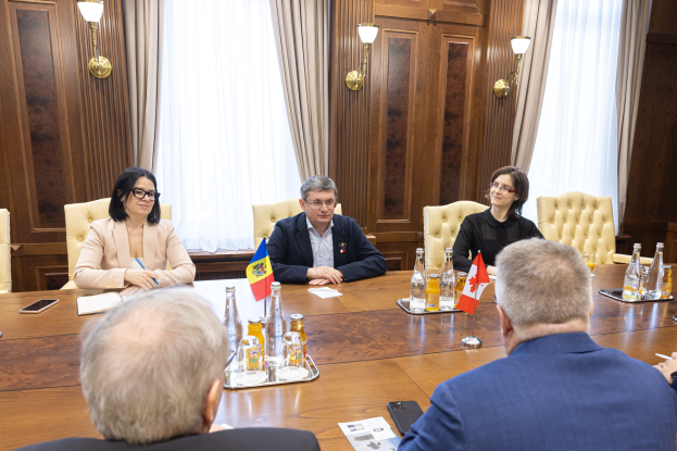 Eine Gruppe von Menschen sitzt um einen Holztisch in einer formellen Umgebung, diskutiert mit EU-bezogenen Dokumenten und Gegenständen, darunter Flaggen, Flaschen, Gläser und Mobiltelefone.