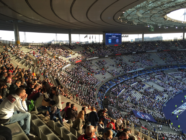 Große Menschenmenge in einem Stadion bei einem Fußballspiel, mit einer Bühne und ein paar Leuten rechts, Fahnen, Stangen, einem Bildschirm und der Allianz Arena in München, Deutschland im Hintergrund.