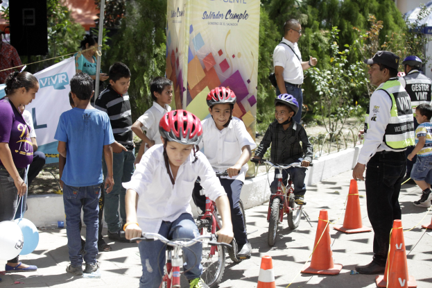Kinder fahren mit Fahrrädern auf einer Straße mit Verkehrskegeln, einige tragen Helme, andere stehen daneben, mit einem Banner, Bäumen und Gebäuden im Hintergrund.