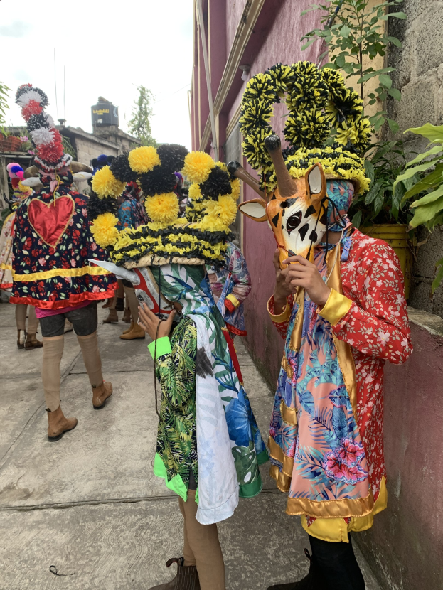 Eine Gruppe von Menschen in bunten Kostümen mit verschiedenen Gegenständen, die vor einem Gebäude während eines Faschingsumzugs mit Bäumen, Gebäuden und einem klaren blauen Himmel im Hintergrund stehen.