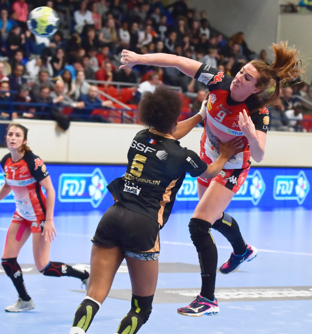 Eine Gruppe von Frauen, die auf einem Platz Handball spielen, mit einem Ball in der Luft, in einem Stadion mit Zuschauern im Hintergrund.