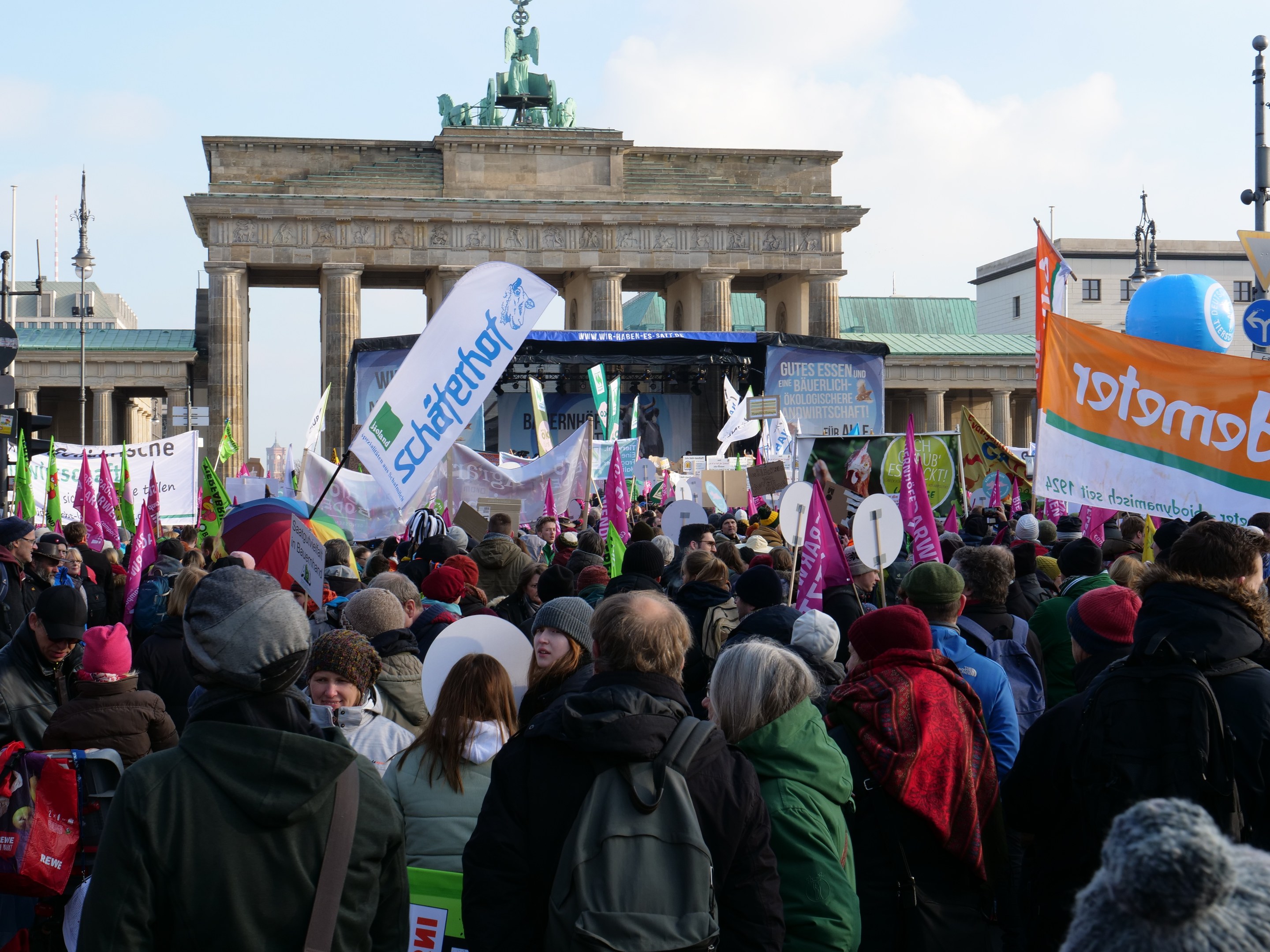 Eine gro├če Menge steht vor dem Brandenburger Tor in Berlin, viele tragen Mützen und haben Taschen dabei, einige halten Schilder; eine Bühne und Laternenmasten sind im Hintergrund zu sehen, der Himmel ist bew├Âlkt.