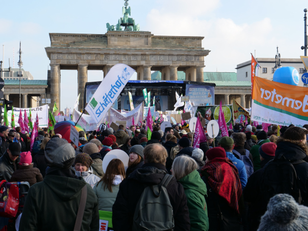 Eine gro├če Menge steht vor dem Brandenburger Tor in Berlin, viele tragen Mützen und haben Taschen dabei, einige halten Schilder; eine Bühne und Laternenmasten sind im Hintergrund zu sehen, der Himmel ist bew├Âlkt.