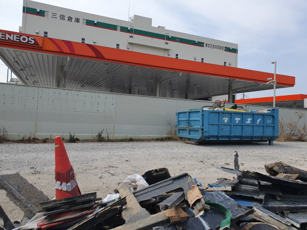 Ein großes Gebäude mit erheblichen Trümmern, darunter Schrott, ein Verkehrskegel und ein blauer Container davor, mit Pflanzen, Polen und dem Himmel im Hintergrund, was auf Abrissarbeiten hinweist.
