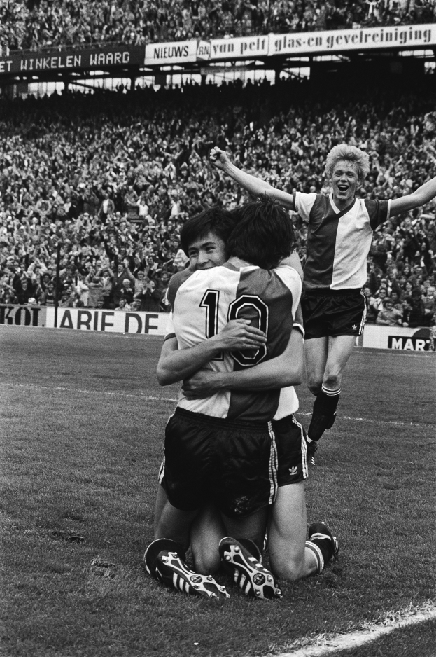 Drei Fussballspieler umarmen sich auf dem Boden und feiern ein Tor, während eine jubelnde Menge und schwenkende Fahnen im Hintergrund zu sehen sind, dargestellt in Schwarz-Weiss.