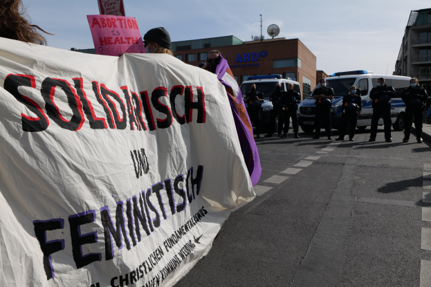 Eine Gruppe von Menschen marschiert mit einem "Solidarität und Feminismus"-Schild die Straße entlang, mit parkenden Fahrzeugen und Gebäuden im Hintergrund unter einem klaren blauen Himmel.