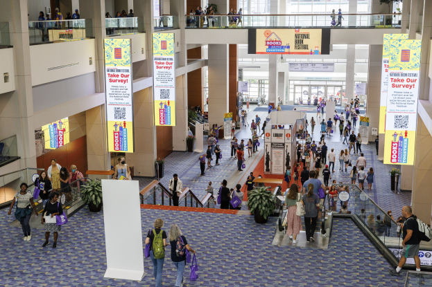 Eine große Gruppe von Menschen, die durch ein großes Gebäude mit Bannern, Topfpflanzen, Rolltreppen, Geländern, Säulen, bedruckten Schildern, Glastüren und anderen Gegenständen geht, einige tragen Taschen, wahrscheinlich nehmen sie am Nationalen Buchfest teil.