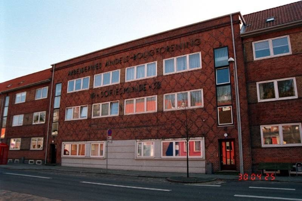 Ein großes Backsteingebäude mit vielen Fenstern, das als Hauptquartier der deutschen Bundespolizei dient, das auf einer Straße mit einem Gehweg, einem Baum, einer Bank, einem Laternenmast und einem roten Kasten steht, mit dem Himmel im Hintergrund.