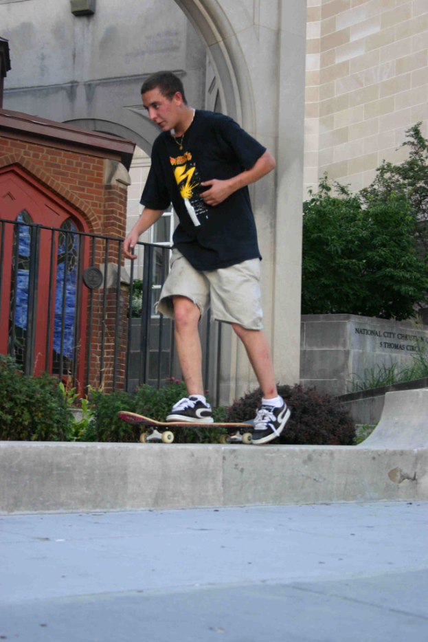 Ein Mann fährt Skateboard auf einer vertikalen Wand mit einer Plattform unten, ein Haus, Grill, Pflanzen, Bäume, eine Ziegelwand und eine Säule im Hintergrund.