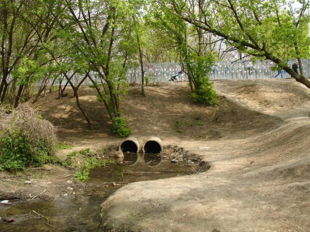 Ein Dirtbike-Park mit einem kleinen Bach, Bäumen und Pflanzen, der eine Graffiti-beschmierte Wand im Hintergrund, einige Menschen und zwei Rohre im Vordergrund unter einem sichtbaren Himmel zeigt.