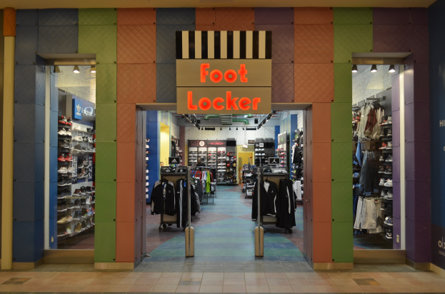 Innenansicht eines Foot Locker Schuhgeschäfts mit Kleidung an Bügeln und Gestellen unter Geschäftsbeleuchtung, mit sichtbarem Boden.
