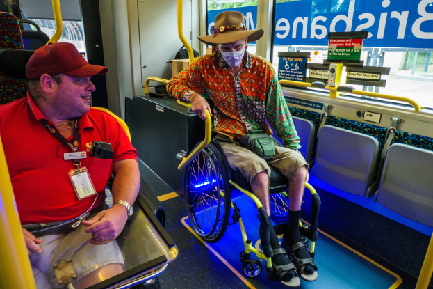 Zwei Männer im Rollstuhl in einem Bus, einer trägt ein rotes T-Shirt und eine Kappe, der andere ein orangefarbenes Hemd und einen braunen Hut, beide tragen Masken und Ausweise, mit gelben Stangen und Glasfenstern, die Bäume, Gebäude und den Himmel zeigen.