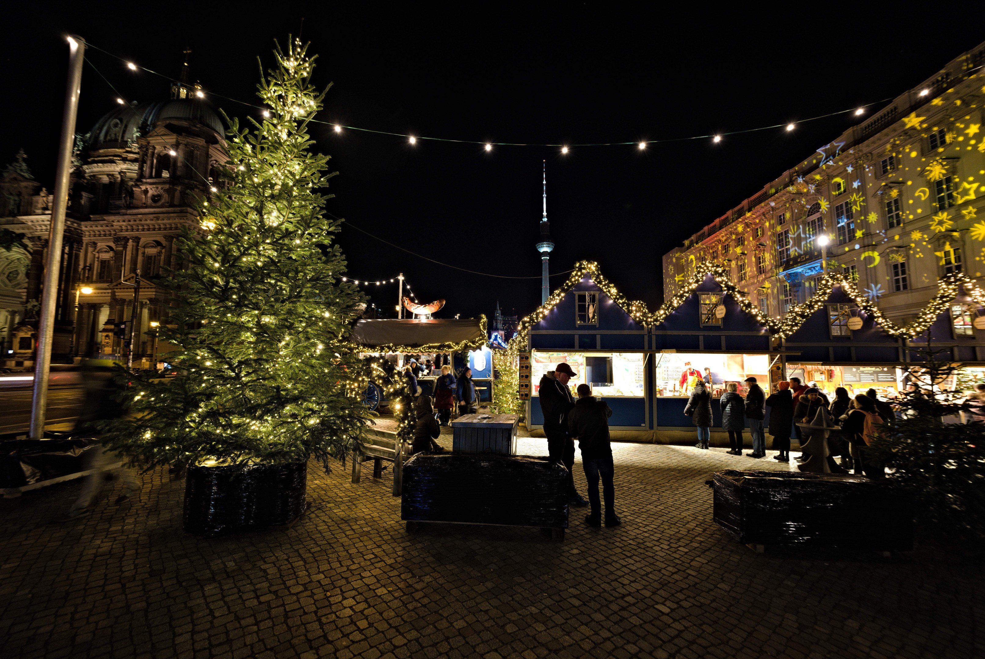 Ein grosser, geschmückter Weihnachtsbaum steht auf einem Berliner Platz, umgeben von Menschen, mit Gebäuden und einer beleuchteten Stange im Hintergrund unter einem dunklen Himmel.