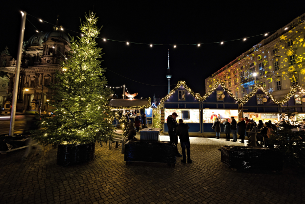 Ein grosser, geschmückter Weihnachtsbaum steht auf einem Berliner Platz, umgeben von Menschen, mit Gebäuden und einer beleuchteten Stange im Hintergrund unter einem dunklen Himmel.
