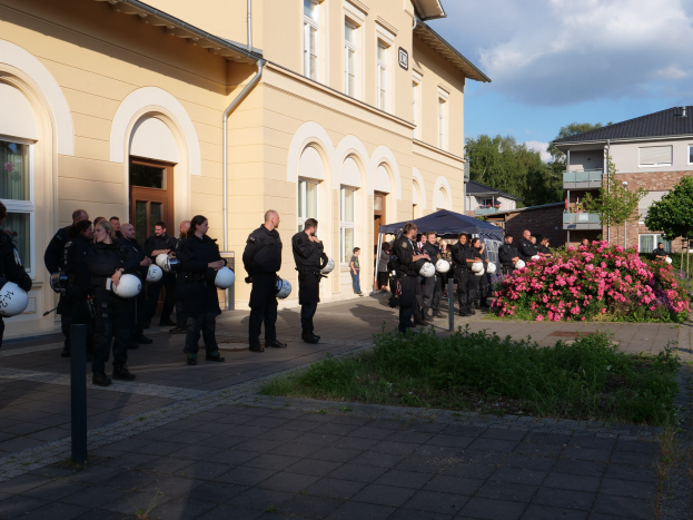 Eine Gruppe von Polizisten vor einem Gebäude mit einem Zelt in der Nähe, umgeben von Grün und einem bewölkten Himmel.